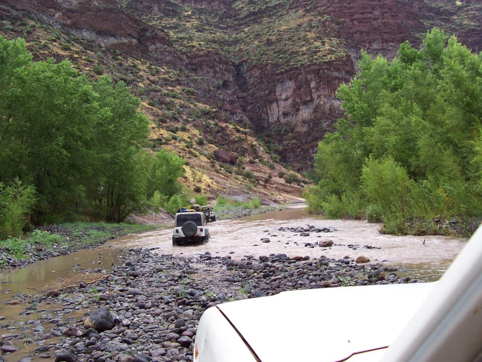 Jeep crossing a rocky river through a red-rock canyon, hood of follow vehicle visible