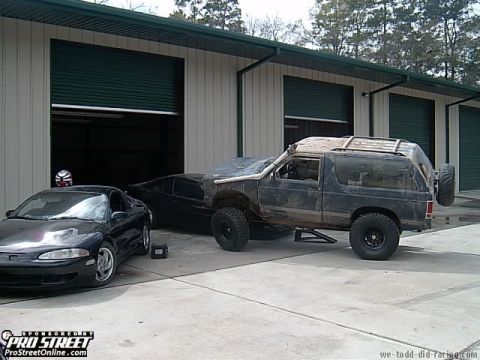 1992 Chevrolet S10 - Modified SUV with large tires and roof rack is parked next to a black coupe in front of a garage.