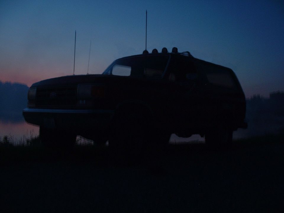Dark silhouette of a full-size truck/SUV with roof lights and antennas against a dusk sky by water