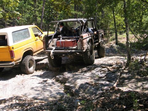 1995 Ford Bronco - Two off-road vehicles navigate a challenging rocky trail through a wooded area.