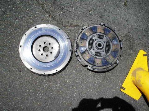 1991 Ford Taurus - A clutch disc and flywheel are laid out on a paved surface, with a yellow object partially visible on the right.