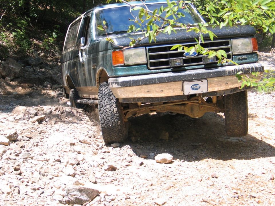 Green Ford Bronco with FSB license plate driving on a rocky trail, branches obscuring the view