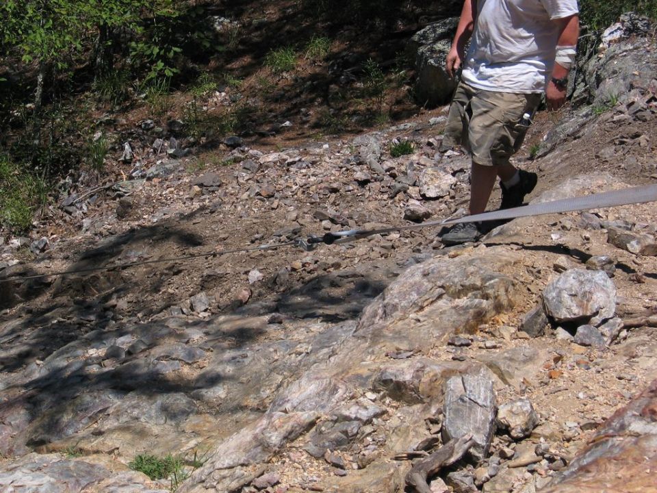 Person in shorts and t-shirt walking across a rocky, wooded slope with a cable stretched across the ground