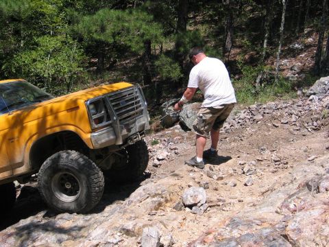1995 Ford Bronco - A man moves a rock from the path of a yellow off-road truck on a trail.