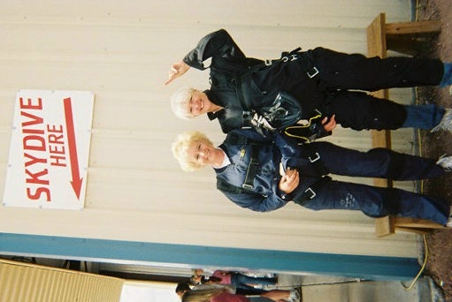 Two people in skydiving harnesses posing next to a SKYDIVE HERE sign