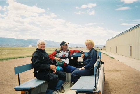 1972 Ford Ranchero - Three individuals in skydiving gear are seated in the bed of a pickup truck, with a large building and mountains in the background.