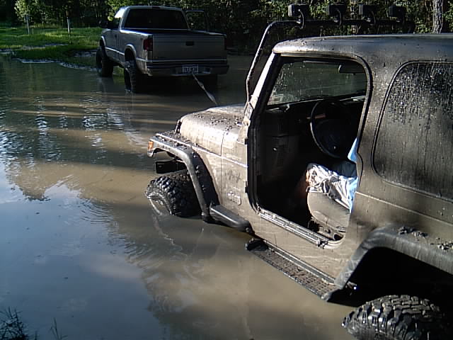 Muddy tan Wrangler in a flooded lot with a gray pickup parked behind in standing water