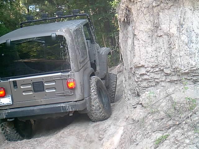 Muddy tan Jeep Wrangler hardtop climbing a rutted dirt trail next to an eroded clay bank