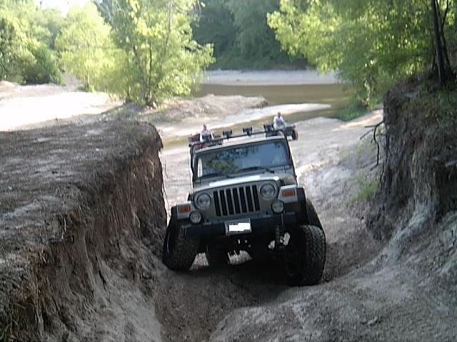 Jeep Wrangler with roof lights climbing a steep rutted dirt trail near a creek