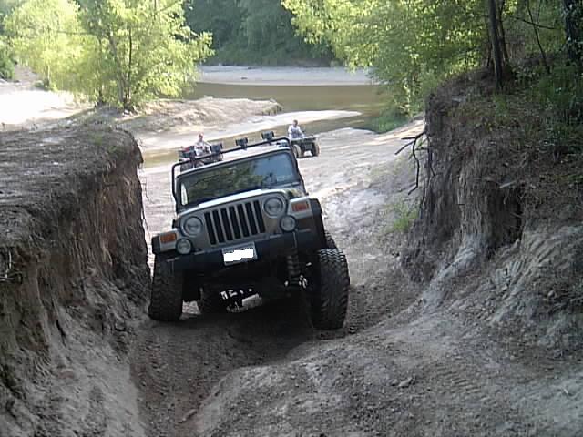 Lifted Jeep Wrangler with light bar climbing a steep sandy off-road trail near a creek