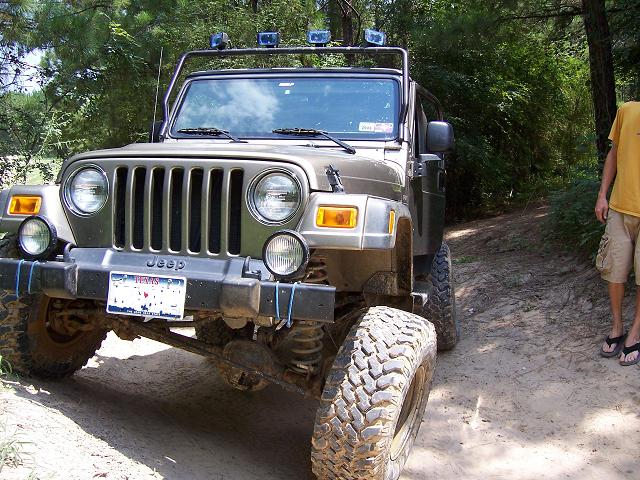 Olive-green Jeep Wrangler with roof lights on a dirt trail, man in yellow shirt at right