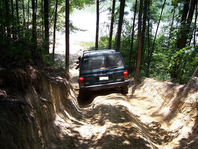 Rear of green Jeep Cherokee XJ crawling a deep sand rut toward a lake through pine trees
