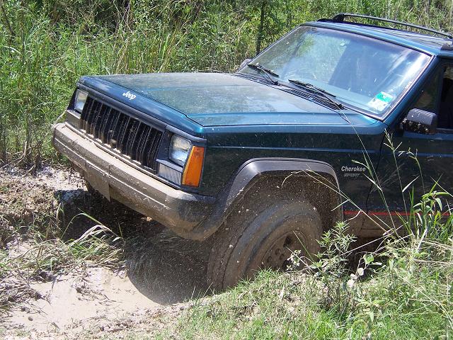 Green Jeep Cherokee Sport stuck in a mudhole, front end covered in mud, tall grass around it