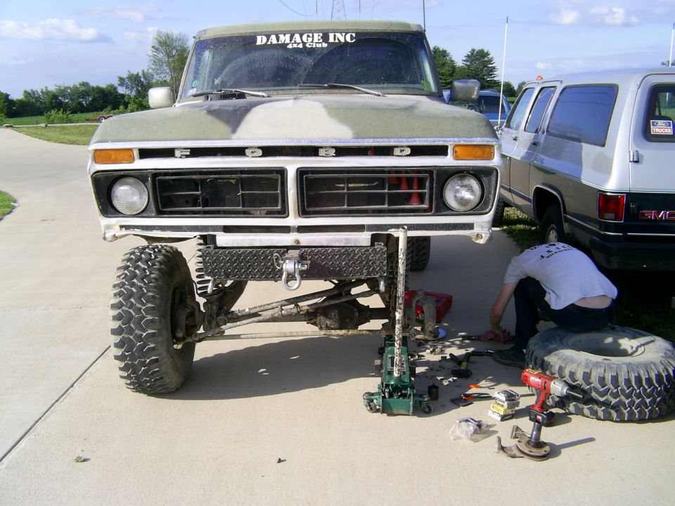 Front view of a lifted 1977 Ford F-150 with camouflage paint, being worked on with a jack and tools. Another SUV is parked nearby