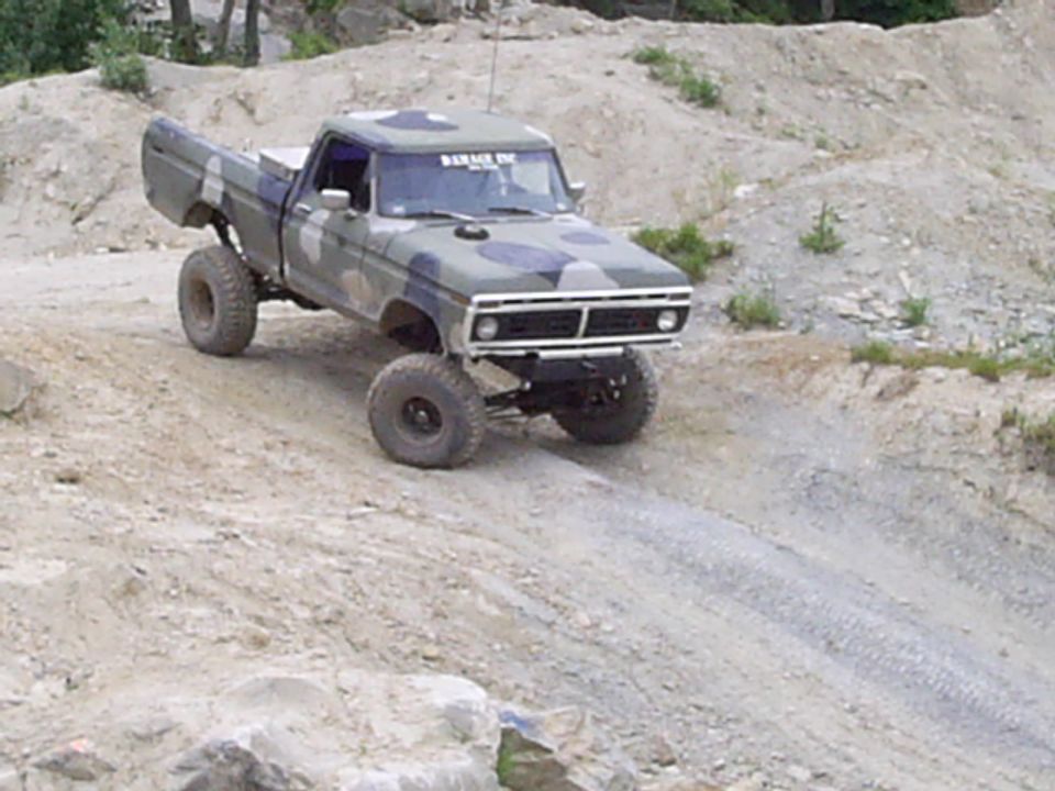 1977 Ford F-150 pickup truck with camouflage paint, lifted suspension, and large tires driving on a dirt hill