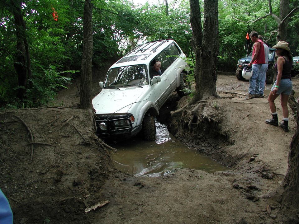 White SUV driving through a muddy ditch with trees around. Another SUV and people are visible in the background