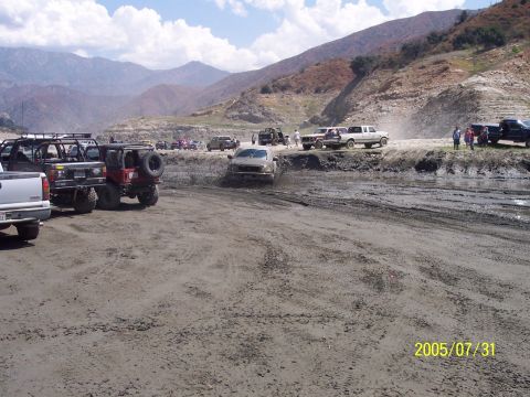 1985 Ford Bronco - Off-road vehicles navigate a muddy terrain under a partly cloudy sky.