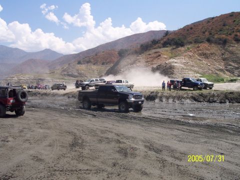 1985 Ford Bronco - A group of off-road vehicles are gathered on a dusty, muddy terrain with mountains in the distance.