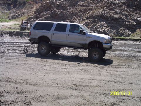 1985 Ford Bronco - A silver SUV with a lifted suspension drives on a dirt road with rocky hills in the background.