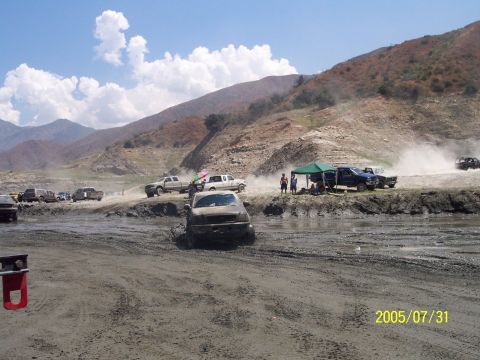 1985 Ford Bronco - Off-road vehicles navigate a muddy terrain under a bright, cloudy sky.
