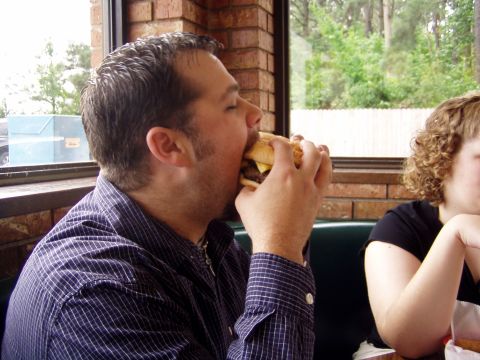 1983 Ford Bronco - Man enjoys a big burger at a restaurant table with a window view.