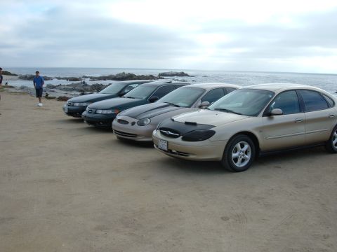 1995 Ford Taurus - Several sedans are parked on a beach, with the ocean and rocky shoreline visible behind them.