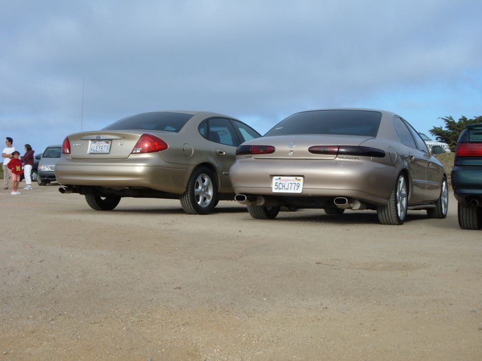 Two tan sedans parked on a dirt lot, rear view. The car on the right has license plate 5CHJ779