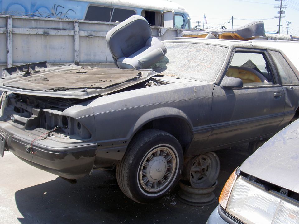 Old car in a junkyard, hood up, seat on top. Another car is next to it