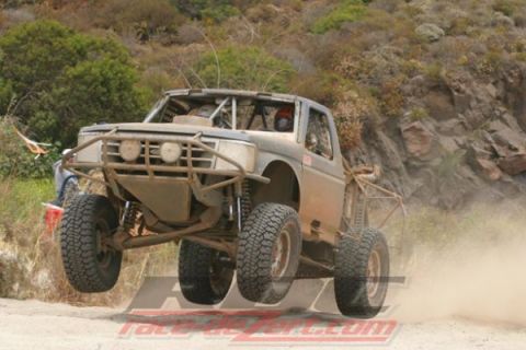 1990 Ford Bronco - tan tube-chassis off-road race truck launches off a jump on a dusty desert course, with a race-dezert.com watermark.