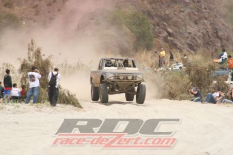 1990 Ford Bronco - off-road race truck flies off a sandy berm as crowds of spectators line the course, with an RDC watermark.