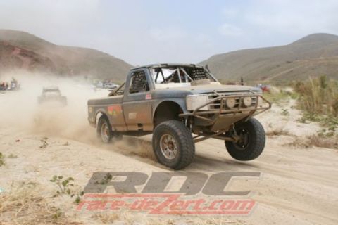 1990 Ford Bronco - tan tube-chassis off-road race truck kicks up dust on a desert dirt course backed by hills, with an RDC watermark.