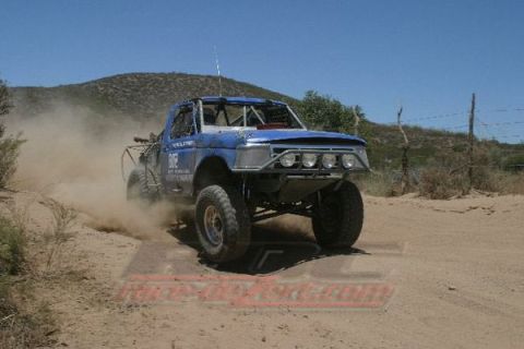 1990 Ford Bronco - blue tube-chassis off-road race truck speeds along a desert dirt road below a hill, with a race-dezert.com watermark.