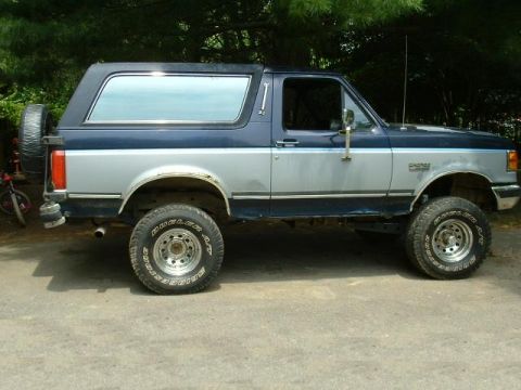 1991 Ford Bronco - A two-tone blue and silver Ford Bronco is parked outdoors, showing its side profile and spare tire.