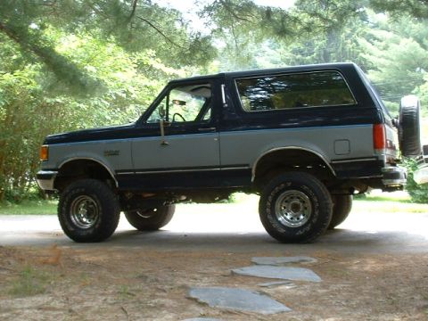 1991 Ford Bronco - This lifted Ford Bronco is shown from the side, parked outdoors on a dirt path with trees in the background.