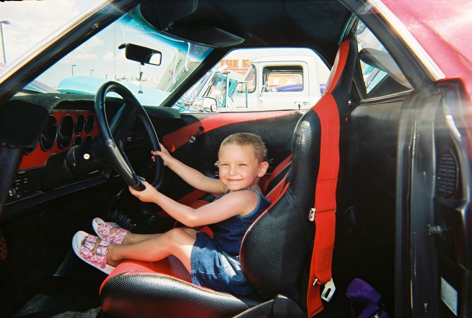 Kid sits behind steering wheel of a car with racing seats. Another truck is visible outside