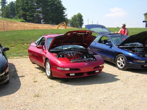 1995 Ford Probe - Red Ford Probe coupe with its hood propped open at an outdoor gravel-lot car meet, showing the transverse V6 engine with a ribbed intake ma…