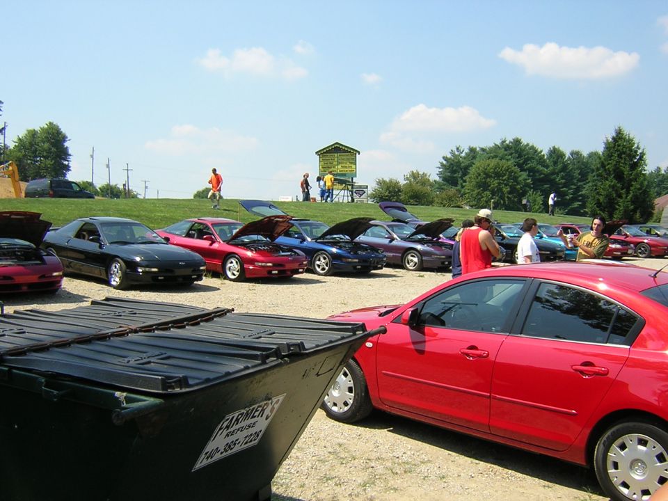 Several cars parked outside with hoods up, people milling around, and a dumpster in the foreground