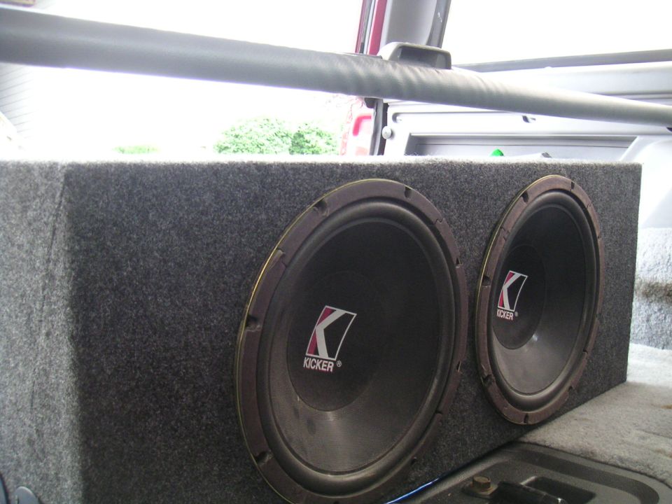Dual Kicker subwoofers in a gray carpeted box, seen from the rear passenger side inside a vehicle