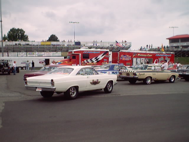 1967 Ford Fairlane and other classic cars parked at a race track with trailers and grandstands in the background