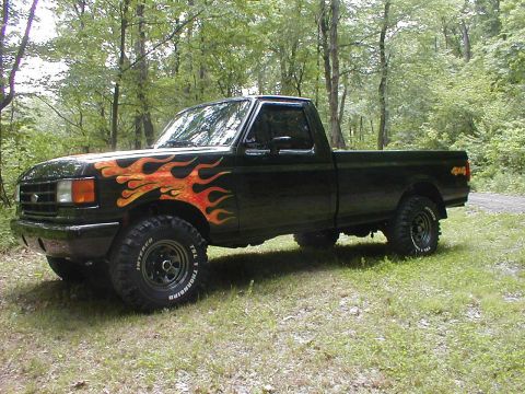 1991 Ford F-150 - Black Ford F-150 regular-cab long-bed pickup with orange and yellow flame graphics on the hood and front fenders, parked on grass at the ed…