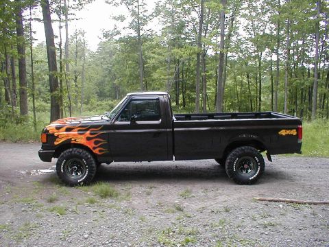 1991 Ford F-150 - A black Ford F-150 pickup truck with flame decals is parked on a gravel road surrounded by trees.