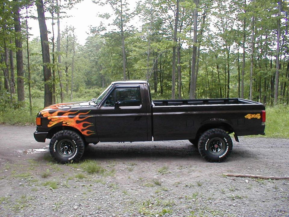 Black Ford F-150 pickup truck with orange and yellow flame decals on the front fender and hood, lifted with off-road tires, parked on a gravel road