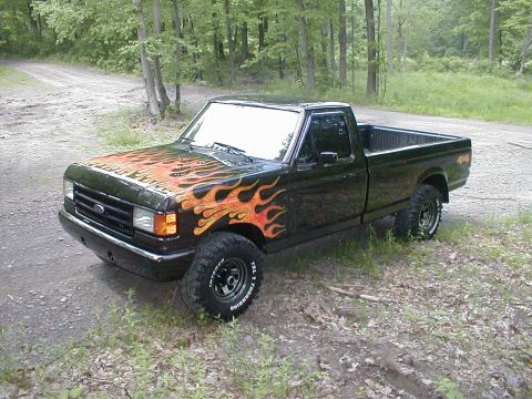 1991 Ford F-150 - A black 1991 Ford F-150 pickup truck with flame decals is parked on a dirt road surrounded by trees.