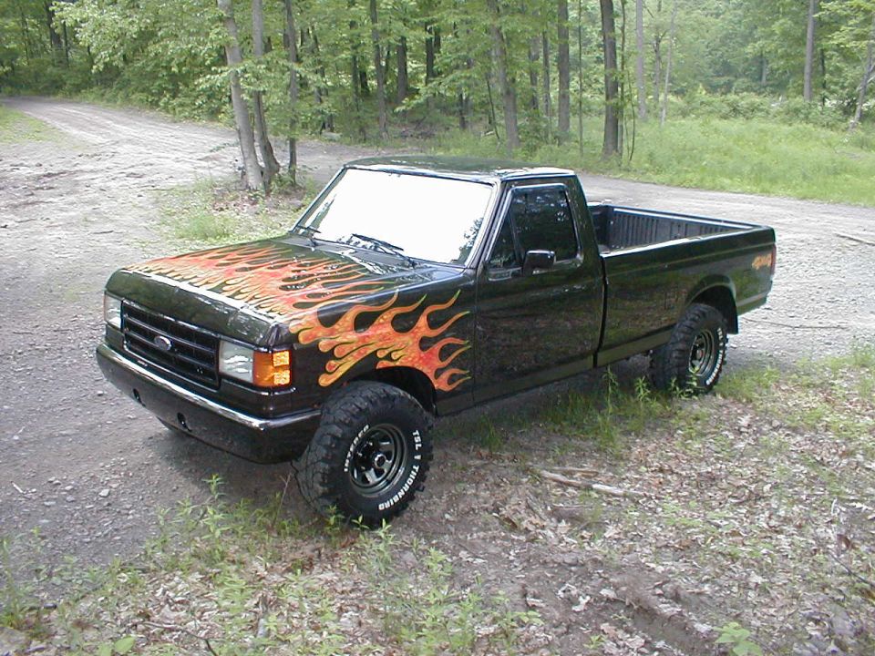Black 1991 Ford F-150 pickup truck with flame decals on the hood and fender, parked on a dirt road