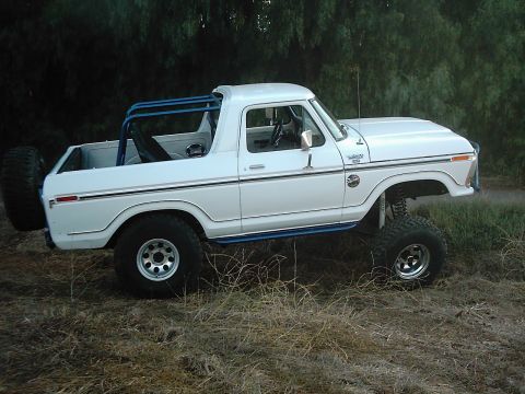 1978 Ford Bronco - A lifted white Ford Bronco with a blue roll cage sits in a grassy, wooded area.