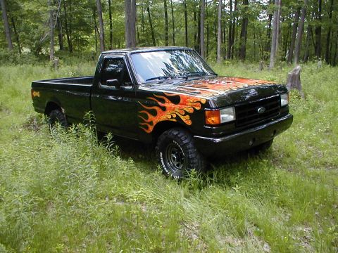 1991 Ford F-150 - This black Ford F-150 pickup truck features flame decals on the hood and front fender, parked in a wooded area.