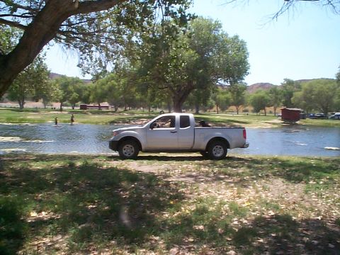2005 Nissan Frontier - A white 2005 Nissan Frontier pickup truck is parked on the grassy shore next to a calm body of water on a sunny day.