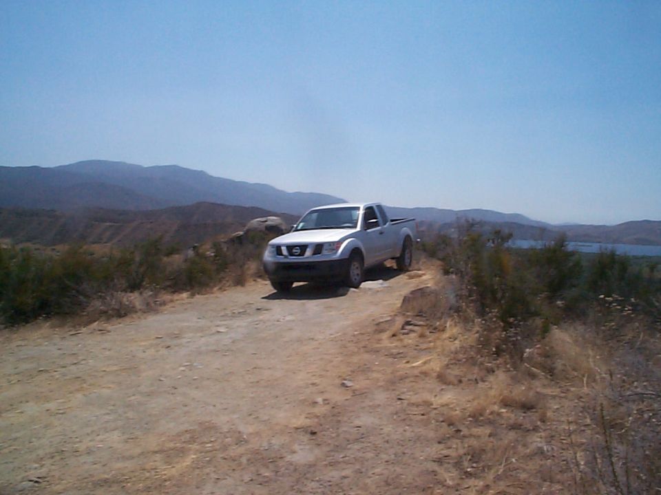 White Nissan Frontier pickup truck on a dirt road with mountains and a lake in the background
