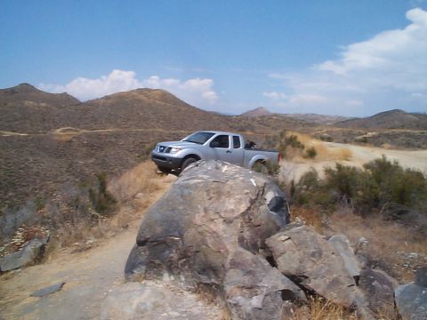 2005 Nissan Frontier - A silver 2005 Nissan Frontier navigates a , off-road path with rocky hills and sparse vegetation under a blue sky.