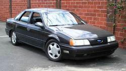 1993 Ford Taurus - A dark sedan is parked in front of a brick wall, with some greenery on the side.